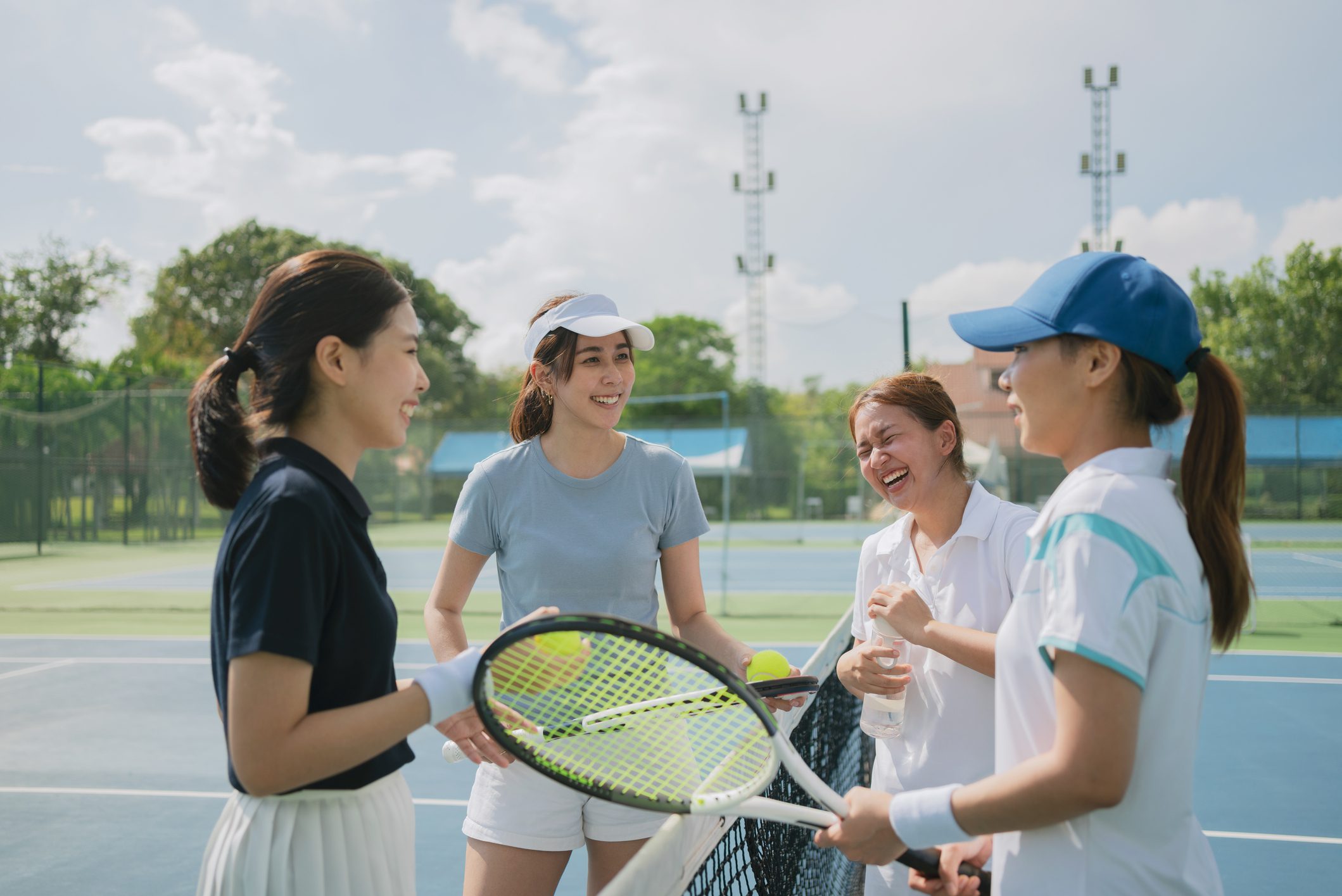 Happy asian young friendship talking and resting after match on the outdoor tennis court on a bright sunny day. Sport activity, tennis training and competition concept
